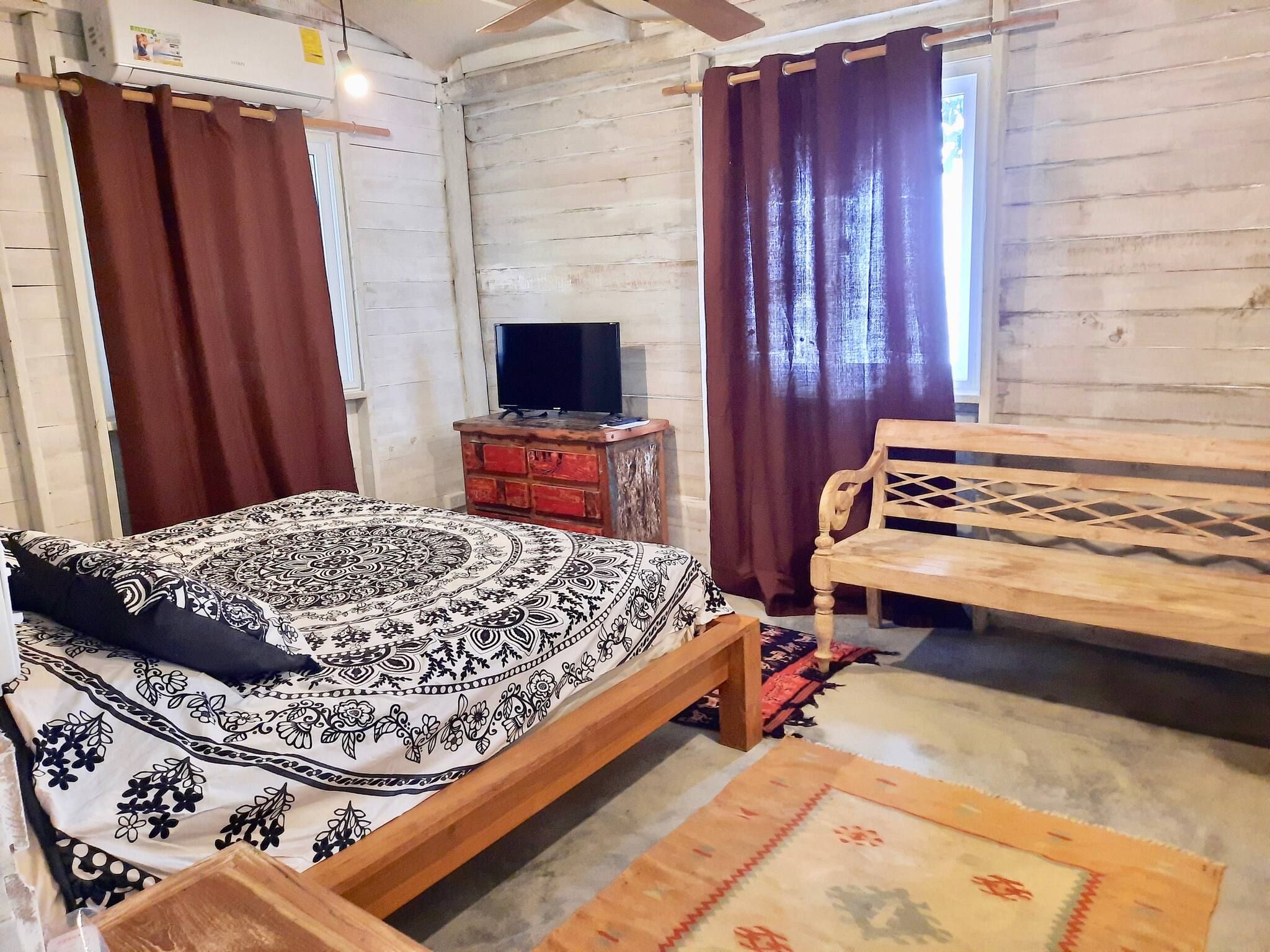 A room with a bed, a wooden bench, a television on a stand, and a window with curtains.