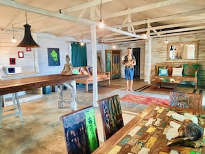 A spacious room with a high ceiling, exposed beams, and a variety of seating options including a sofa, a chair, and a bench. There is a large table with a colorful abstract painting and a sculpture. - Incredible Loft on the Beach! (Bocas del Toro)