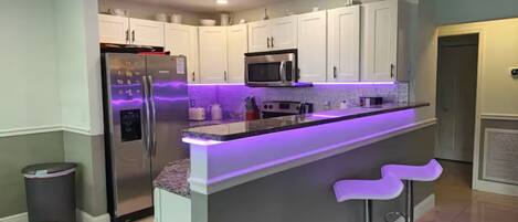 Een moderne keuken met een centraal eiland, ingebouwde apparatuur en paars verlichting onder de keukenkastjes.