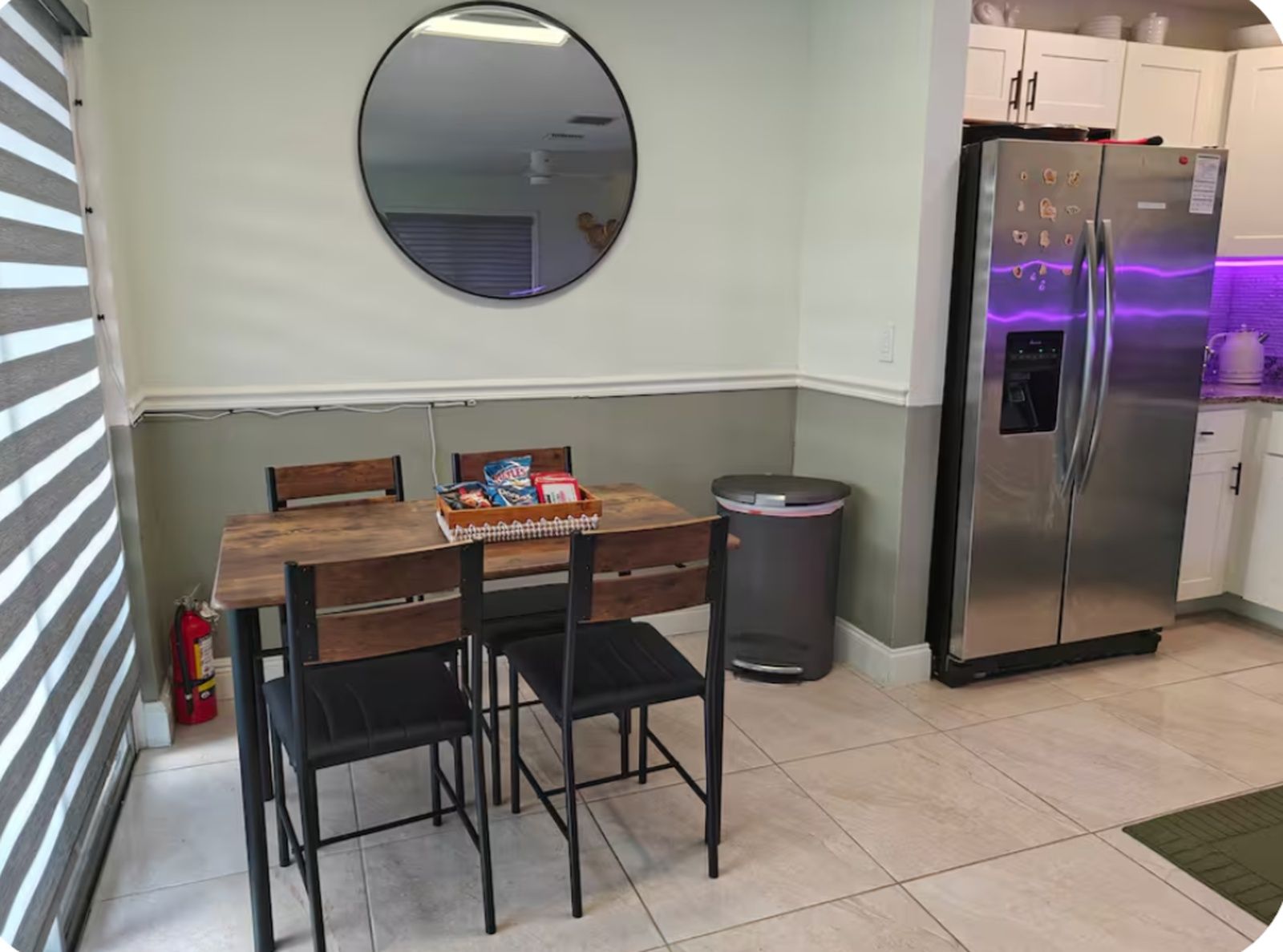 A compact kitchen with a dining table, chairs, a trash bin, and a refrigerator.