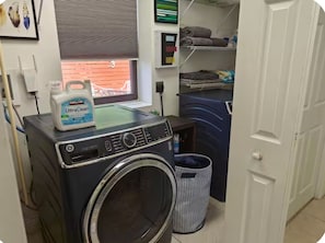 Ein Waschbereich mit einer Frontlader-Waschmaschine, einer Waschmittelflasche, einem Fenster mit Jalousien und einem Regal mit gefalteten Handtüchern.