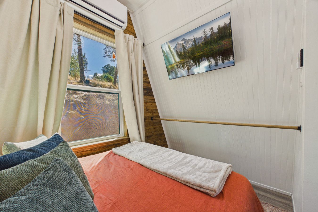 Une chambre à coucher avec un lit, une fenêtre avec des rideaux, un tableau représentant un paysage de montagne et une poutre en bois.