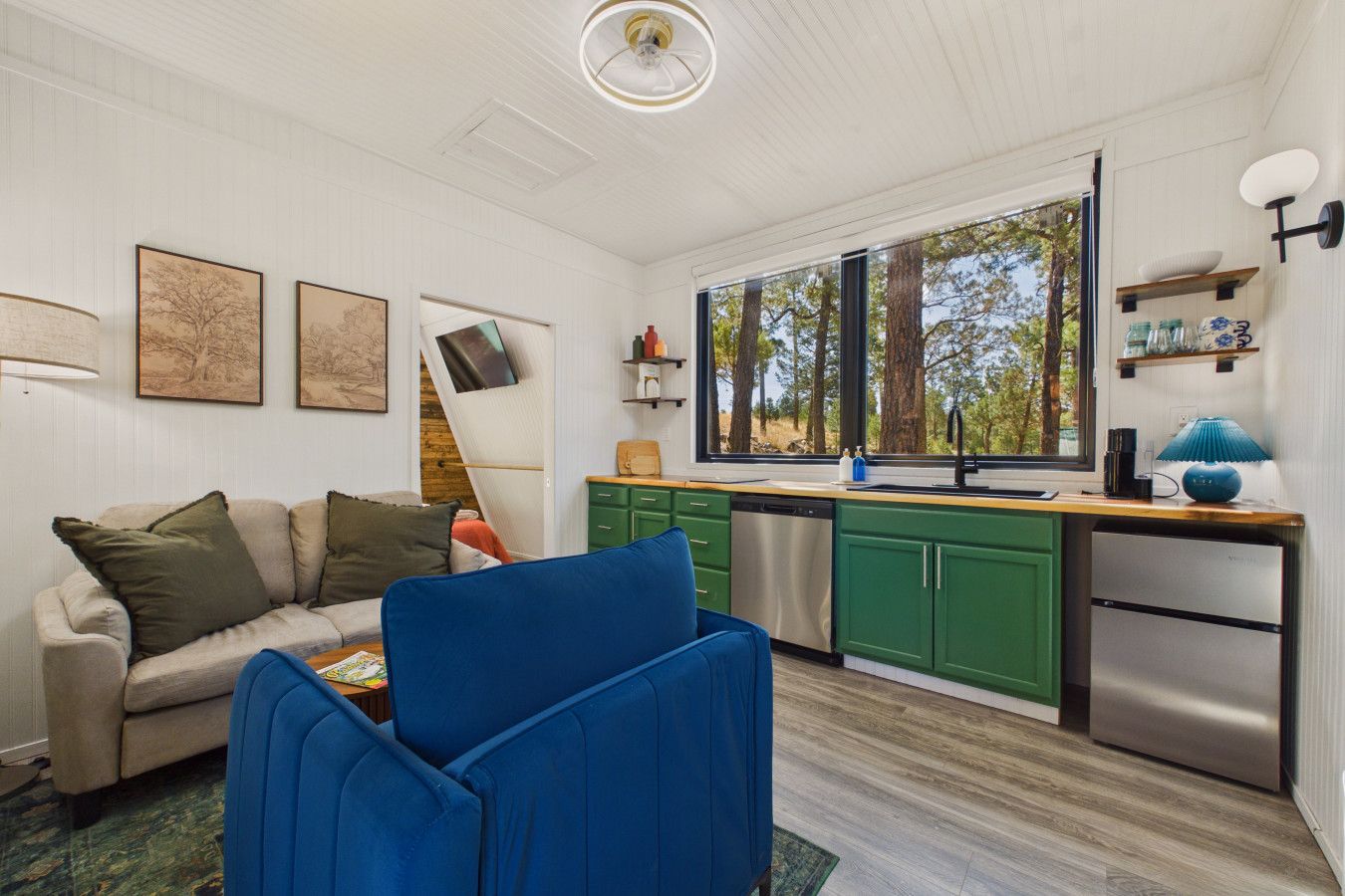 Un espace de vie compact comprenant un canapé, un fauteuil bleu, une cuisine avec des meubles verts et une grande fenêtre donnant sur la verdure.