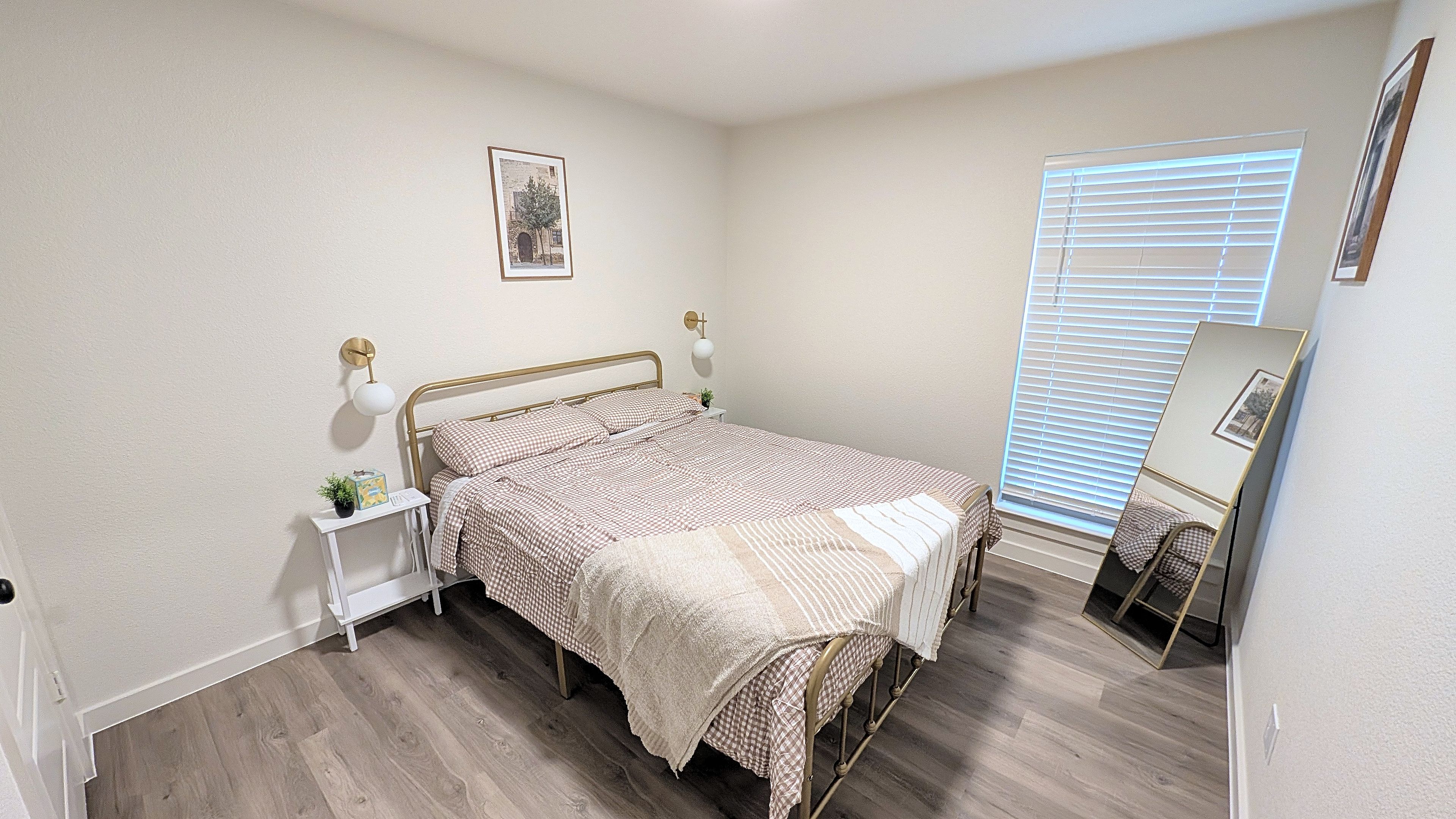 A bedroom with a bed, bedside tables, a wall-mounted lamp, a framed picture, and a window with blinds.
