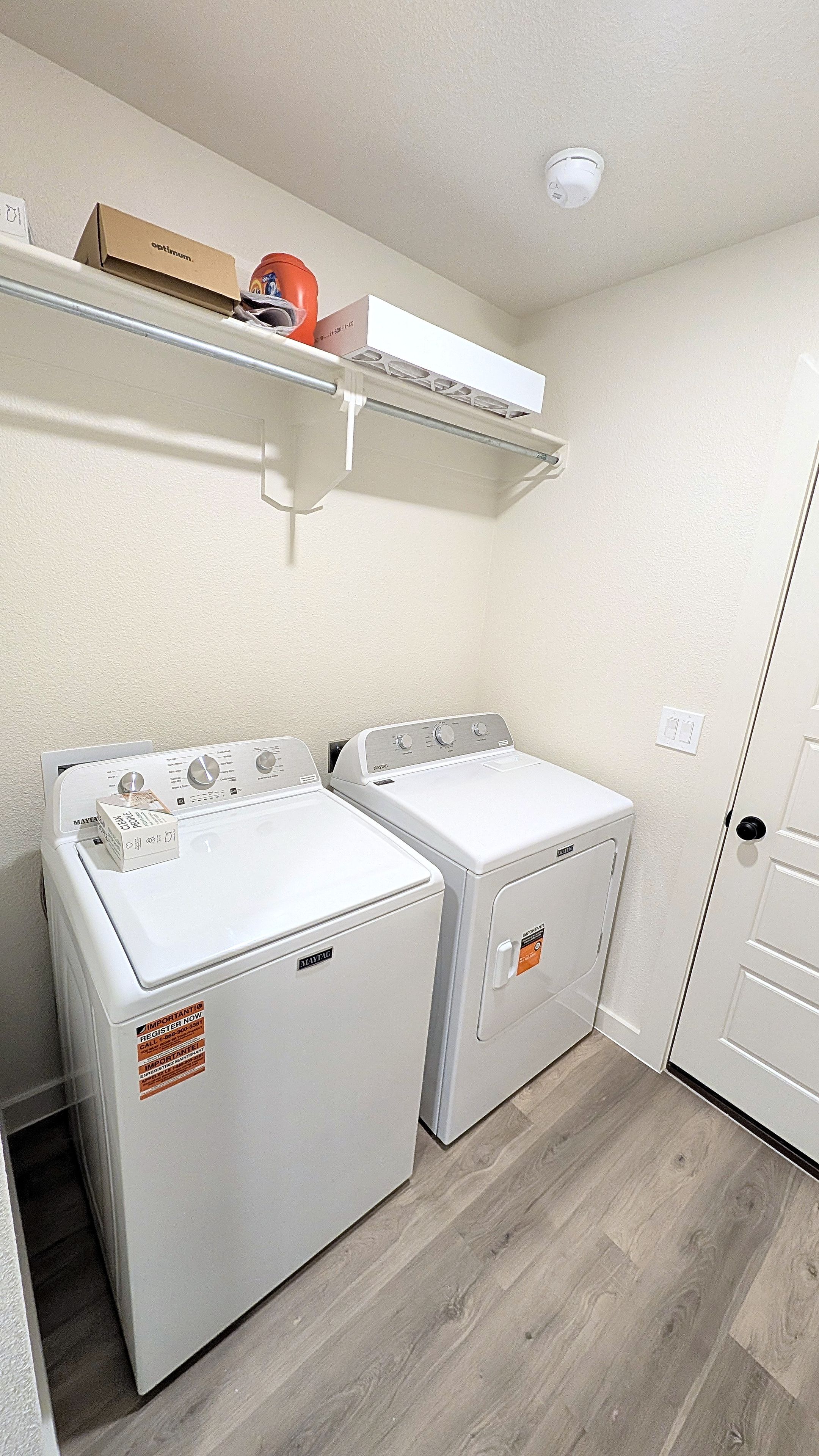 Una sala de bugaderia amb dues rentadores de càrrega frontal, una estant amb una caixa i un objecte vermell, i una porta amb interruptor de llum.