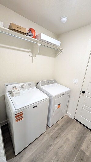 A laundry room with two front-loading washing machines, a shelf with a box and a red item, and a door with a light switch. - Auburn House - New 4BR Near Texas Tech (Lubbock)
