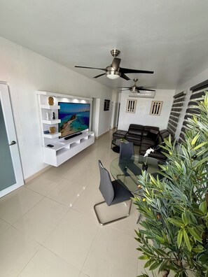 A modern living room with a flat-screen TV, a glass-top dining table, and a ceiling fan. - Villa Valmira | private pool (Canóvanas)