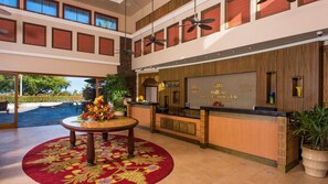 A hotel reception area with a reception desk, a round table with a floral centerpiece, and a view of the outdoor pool area. - Luxury 2BR Plus Suite on Hawaii's Kohala Coast Near Golf & Beaches (Waikoloa Village)