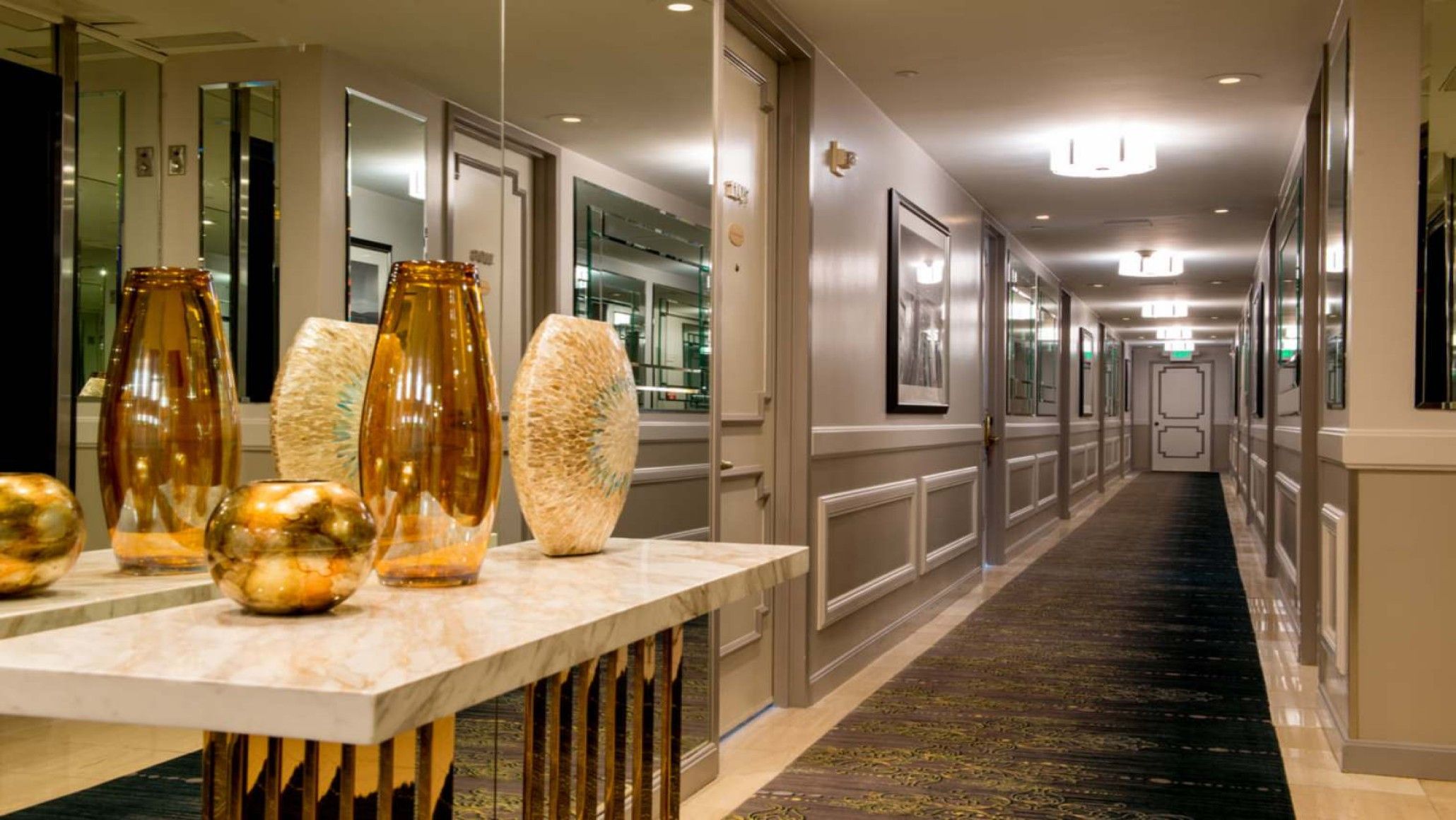 A hotel corridor with a marble reception desk, large vases, and framed artwork on the walls.