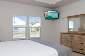 A bedroom with a bed, a wooden dresser, a television mounted on the wall, and a window with blinds. - Sweet 'N Salty (Port St. Joe)