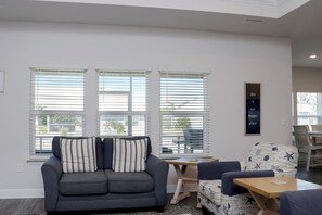 A living room with a sofa, a table, and a chair. There are three windows with blinds, a wall-mounted picture, and a ceiling light. - Sweet 'N Salty (Port St. Joe)