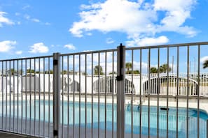 Una piscina con una cerca blanca al frente, un cielo azul despejado y edificios con palmeras al fondo.