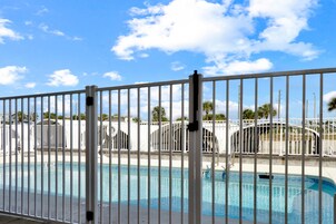 Una piscina con una cerca blanca al frente, un cielo azul despejado y edificios con palmeras al fondo.