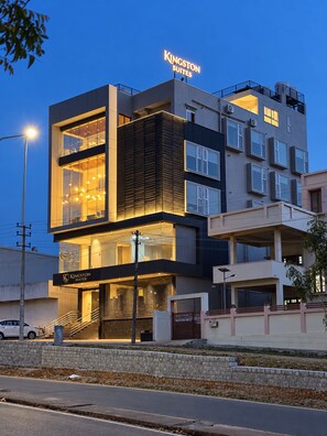 A modern hotel with a prominent sign, multiple floors, and a well-lit entrance. - Kingston Suites (Mysore)