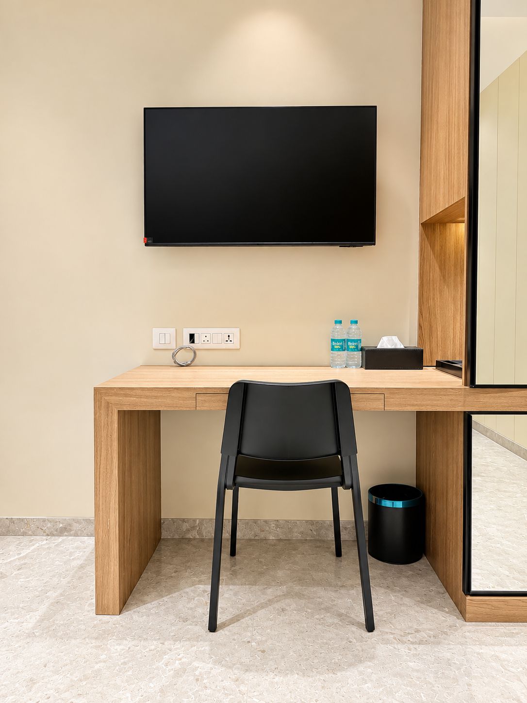 A modern hotel room with a wooden desk, a black chair, a flat-screen TV mounted on the wall, a small table with water bottles, and a trash can.