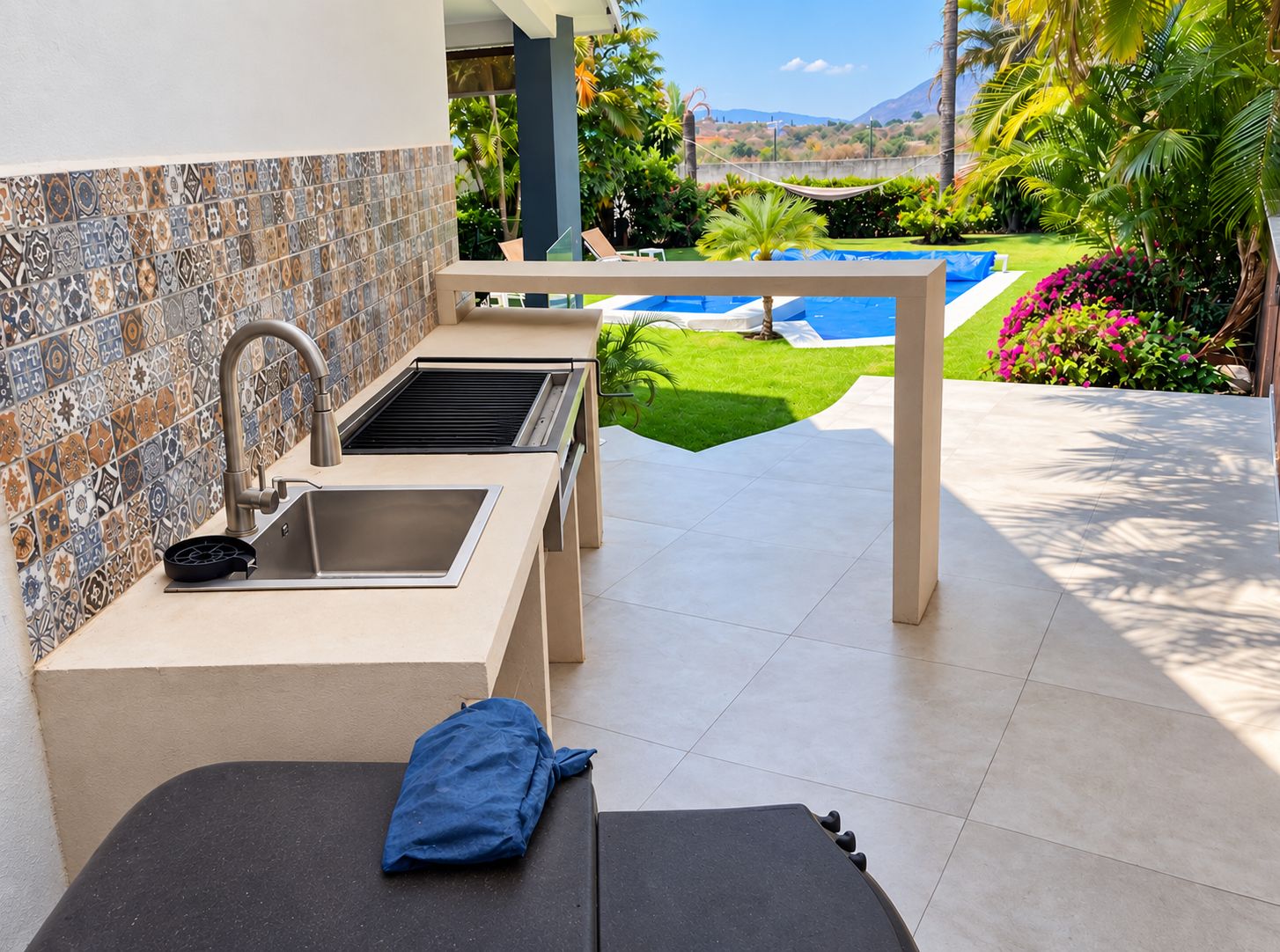 An outdoor kitchen area with a tiled backsplash, a sink, and a grill, overlooking a swimming pool and lush greenery.