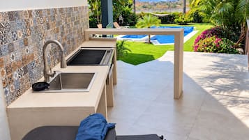 An outdoor kitchen area with a tiled backsplash, a sink, and a grill, overlooking a swimming pool and lush greenery.