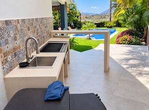 Angolo cucina esterno con schienale piastrellato, lavello e barbecue, con vista su una piscina e vegetazione lussureggiante.