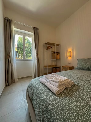 A bedroom with a bed, a window with curtains, a bedside table, and a wall-mounted lamp. - defaultName (Olbia)