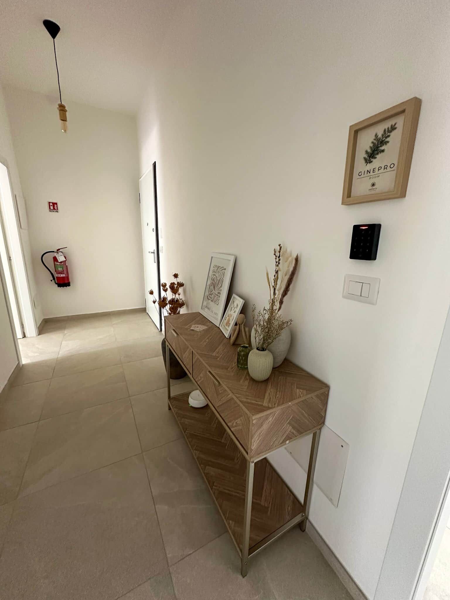 A hallway with a wooden console table, framed artwork, a fire extinguisher, and a wall-mounted light fixture.