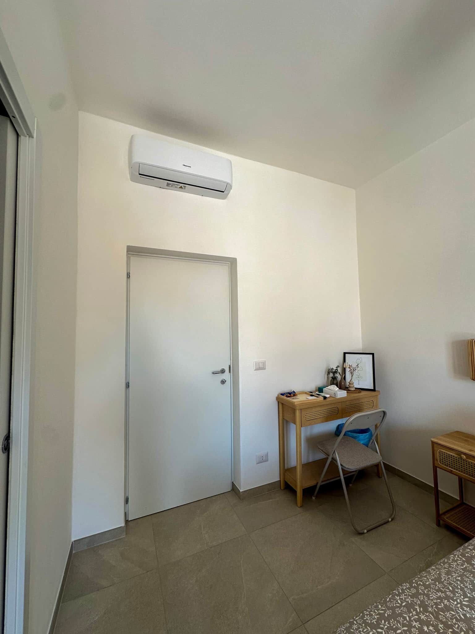 Une pièce avec une porte blanche, un climatiseur fixé au mur, un bureau avec une chaise et une petite table avec une lampe.