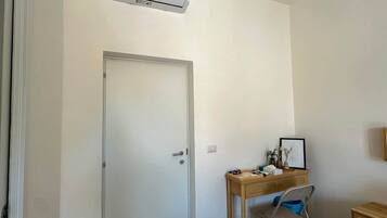 Une pièce avec une porte blanche, un climatiseur fixé au mur, un bureau avec une chaise et une petite table avec une lampe.