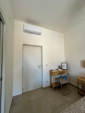 A room with a white door, an air conditioning unit mounted on the wall, a desk with a chair, and a small table with a lamp. - defaultName (Olbia)