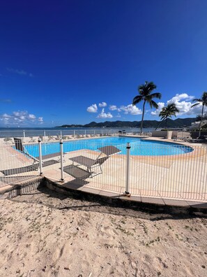 Piscina com cerca branca, rodeada por uma área arenosa com palmeiras.
