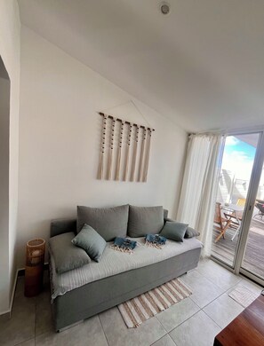 Ein modernes Wohnzimmer mit einem grauen Sofa, einer gewebten Wanddeko und Blick ins Freie durch eine Glastür.