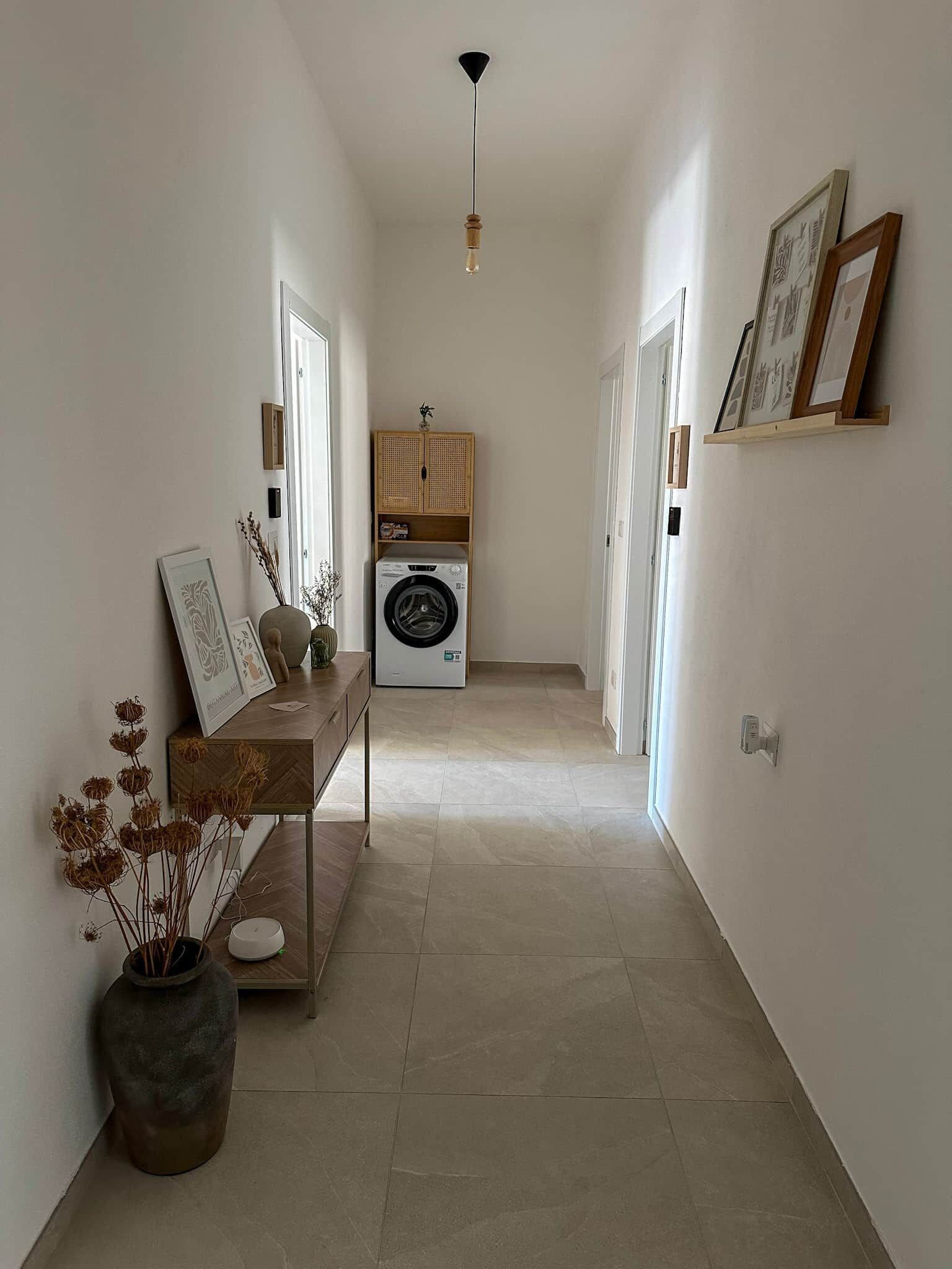 Un corridoio con lavatrice, una panca in legno con oggetti decorativi e quadri appesi alle pareti.