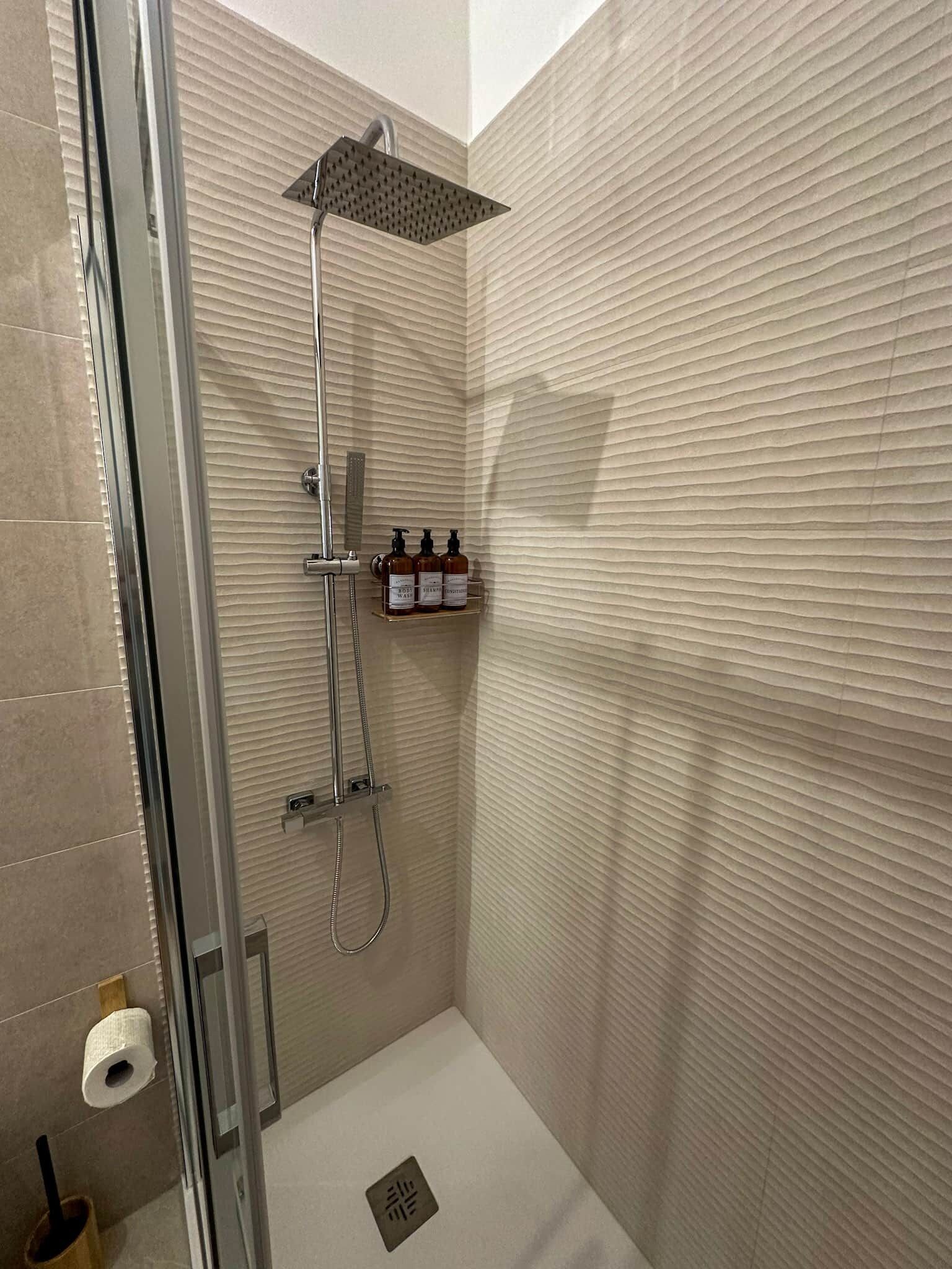 Una ducha con cabezal de ducha manual, un accesorio de ducha manual y un cabezal de ducha instalado en la pared. Hay un estante con artículos de tocador.