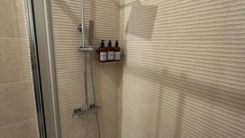 Una ducha con cabezal de ducha manual, un accesorio de ducha manual y un cabezal de ducha instalado en la pared. Hay un estante con artículos de tocador.