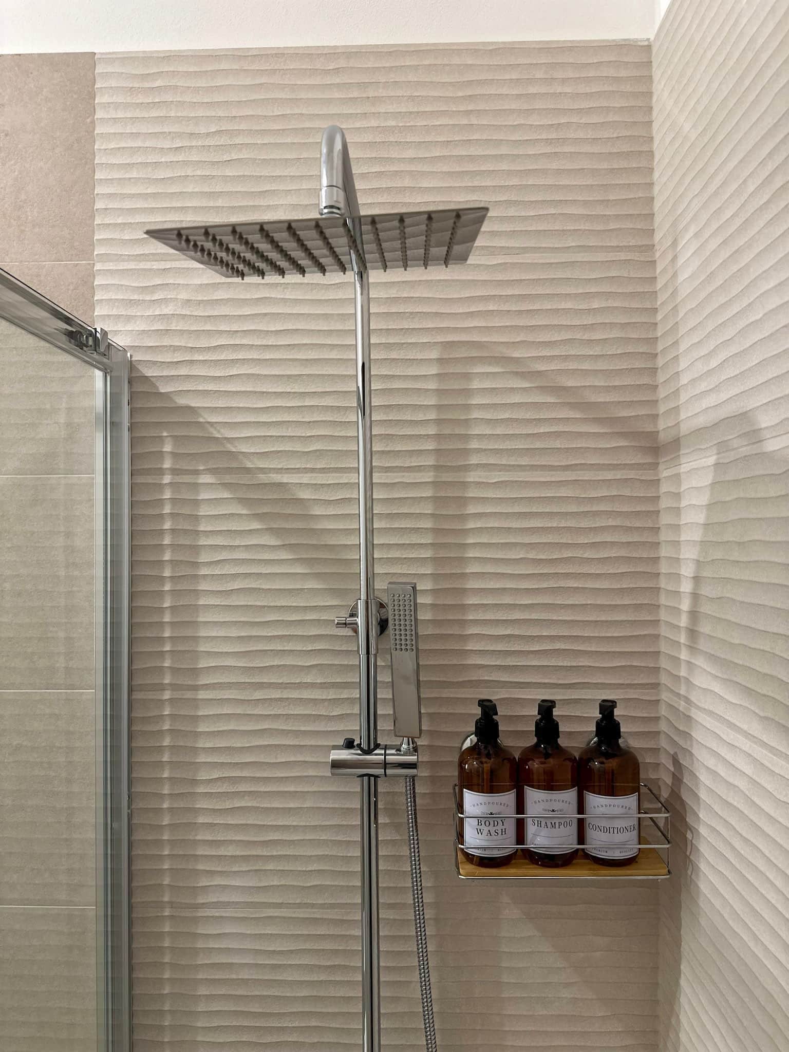 Une douche équipée d’un pommeau de douche à main et d’un panneau de commande fixé au mur. Sur une étagère, on trouve trois flacons de gel douche, de shampoing et d’après-shampoing.