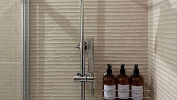 Une douche équipée d’un pommeau de douche à main et d’un panneau de commande fixé au mur. Sur une étagère, on trouve trois flacons de gel douche, de shampoing et d’après-shampoing.