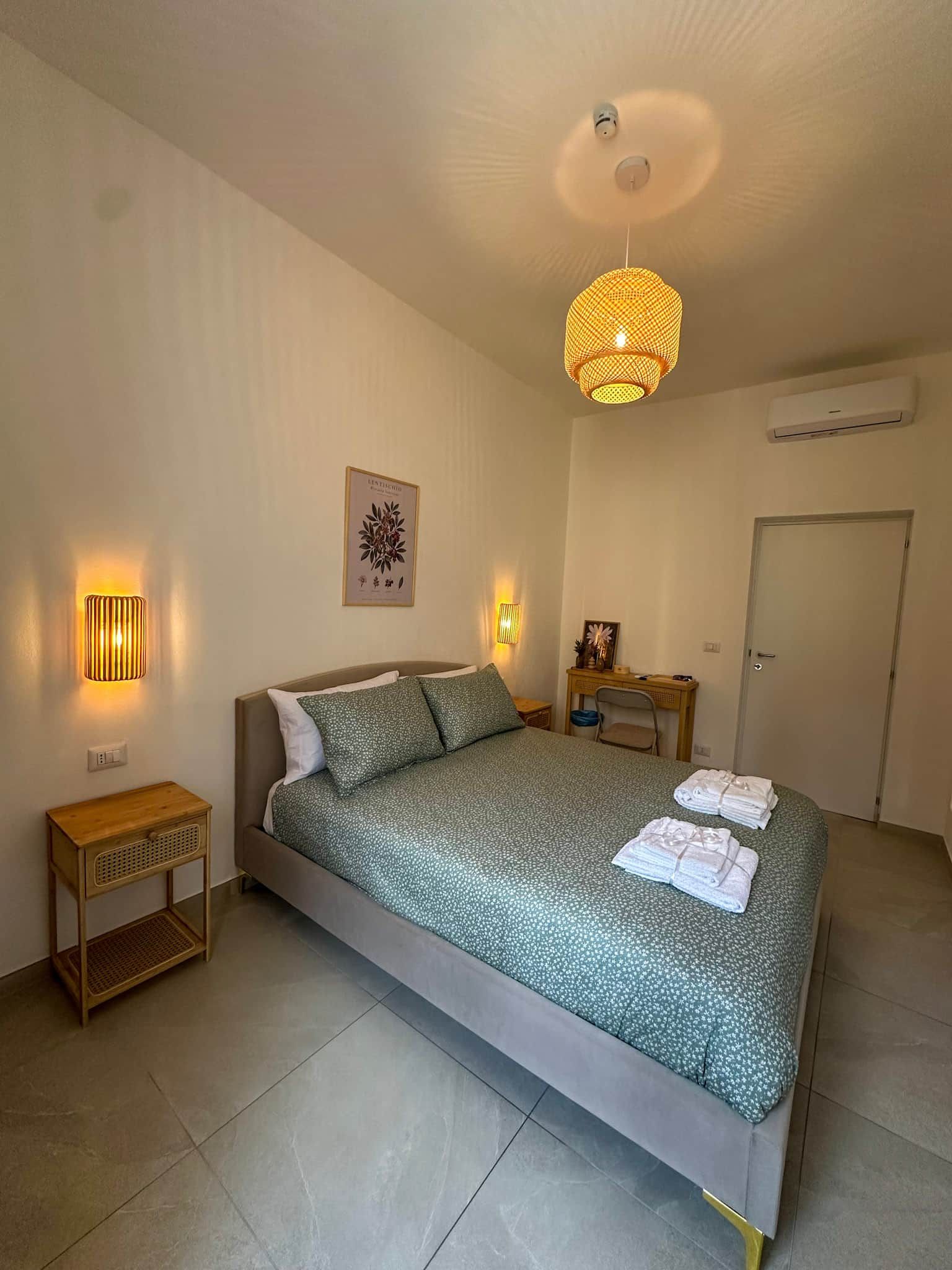 A bedroom with a bed, bedside lamps, a ceiling light, a door, and a wall-mounted air conditioning unit.
