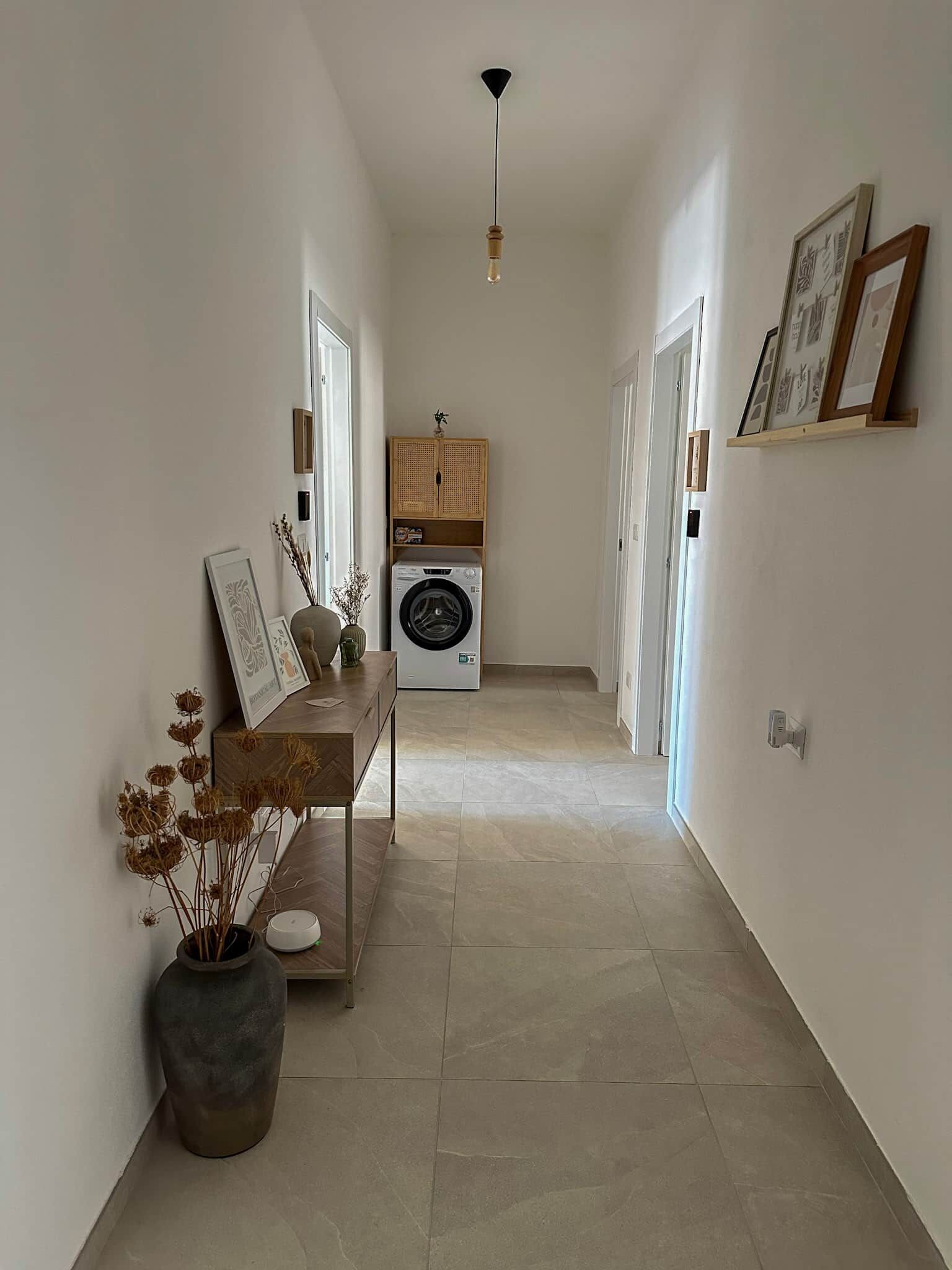 A narrow hallway with a washing machine, a bench with a vase of dried flowers, and framed pictures on the walls.