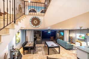 Ampla sala de estar com mesa de bilhar, mesa de jantar e assentos confortáveis. Uma escadaria leva a um andar superior com teto decorativo.