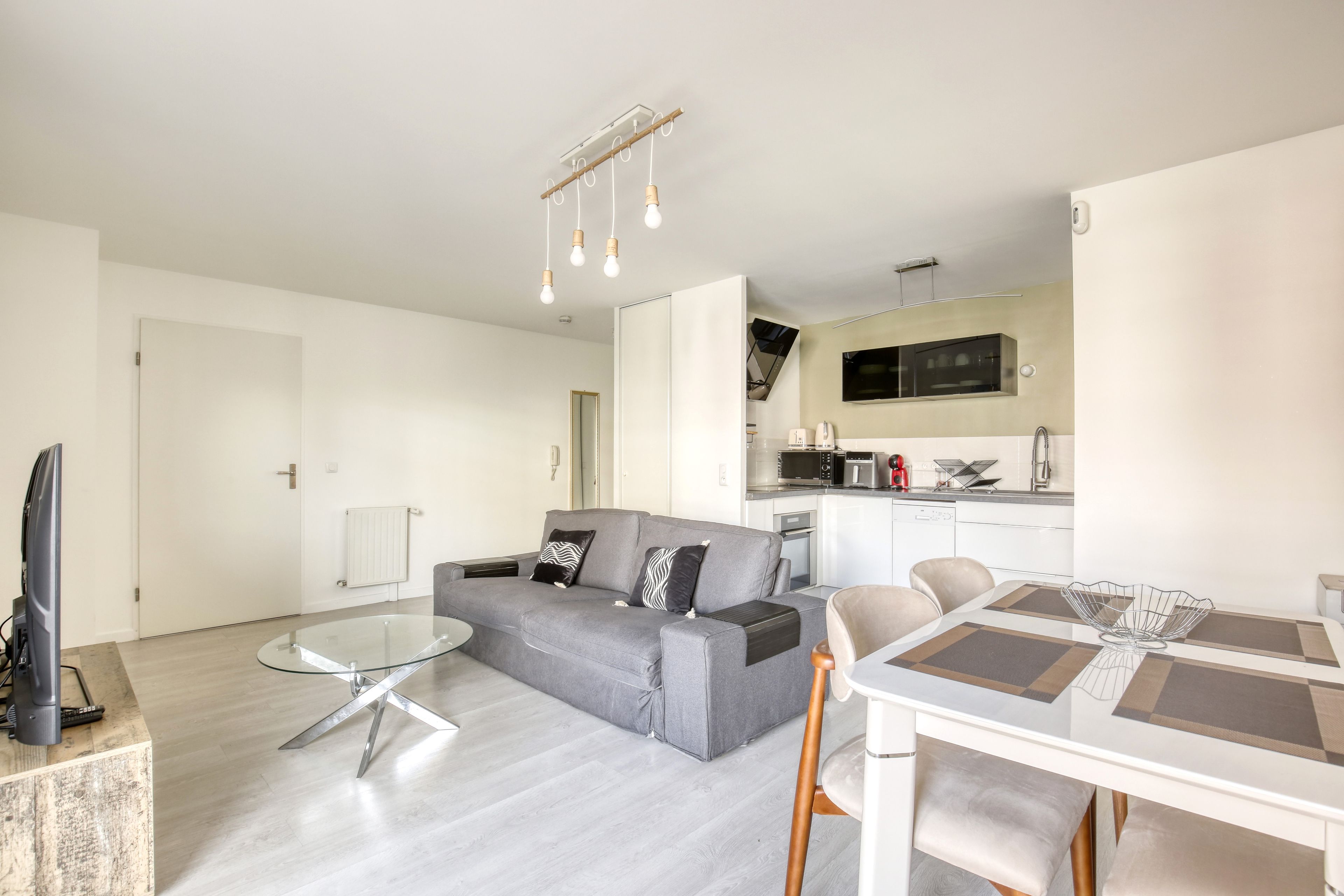 Un salon moderne avec un canapé gris, une table basse en verre et un coin repas comprenant une table et des chaises blanches.