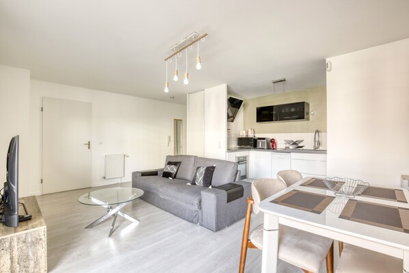 A modern living room with a grey sofa, a glass coffee table, and a dining area with a white table and chairs. - Vrbo Property (Franconville)