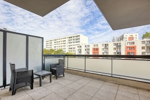 Een balkon met twee rieten stoelen en een klein tafeltje, met uitzicht op een rij meerlaagsgebouwen met balkons.
