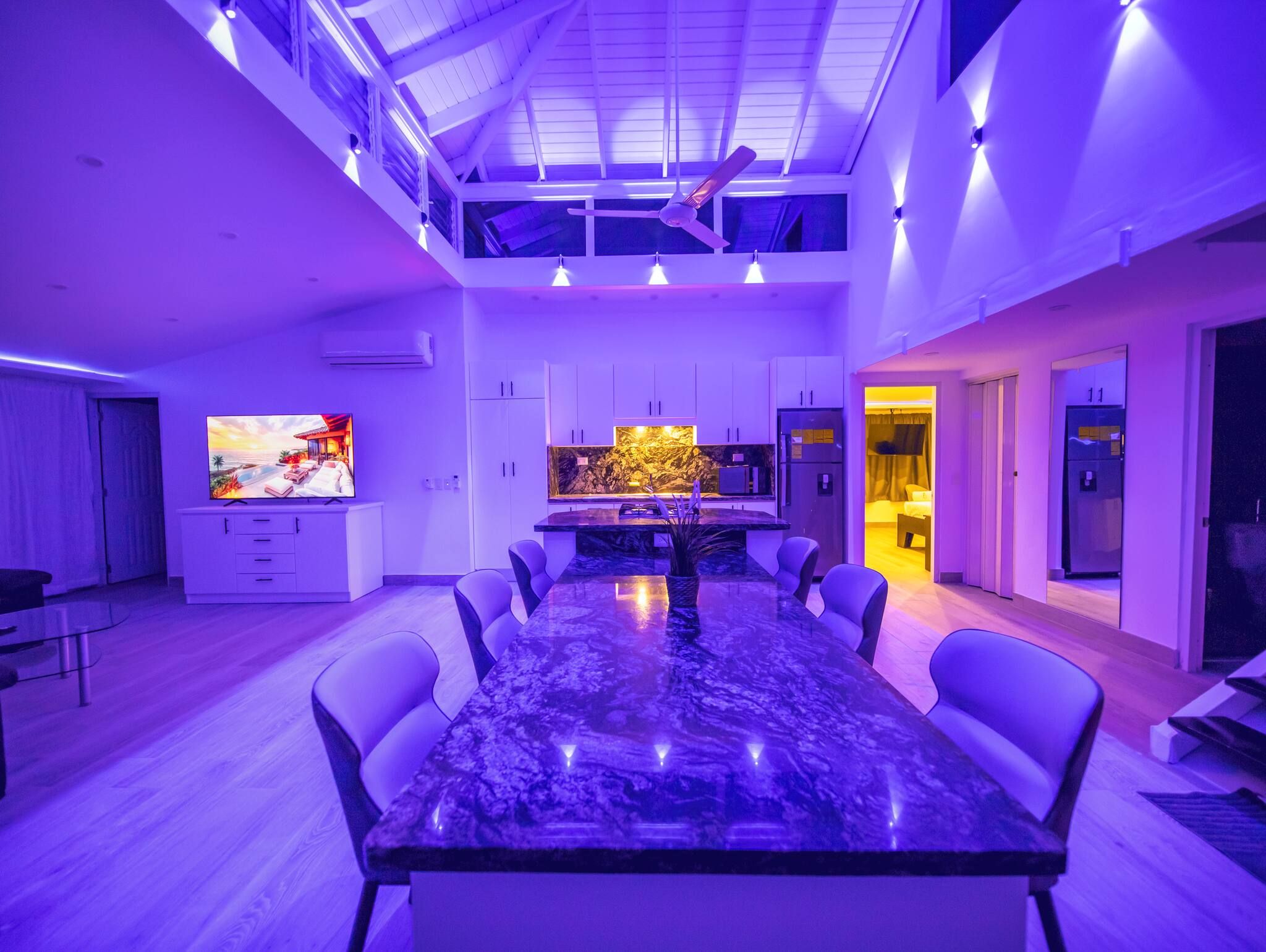 Una cocina moderna con una isla central, un comedor con una mesa grande y sillas, y un televisor que muestra una imagen panorámica.