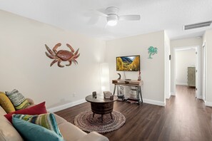 A living room with a ceiling fan, a wooden coffee table, a couch with colorful cushions, a wall-mounted crab decoration, and a painting of a landscape. - Walk to Beach • Heated Pool • 3 King Beds • Boat Ramp • St. Augustine (St. Augustine)