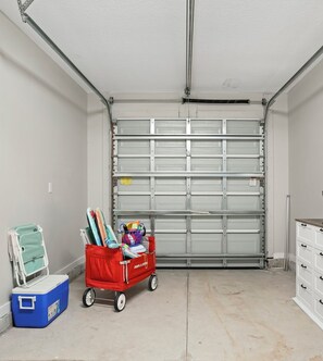 En garage med lukket garagedør, en rød indkøbsvogn med varer, en blå køleboks og en hvid kommode.
