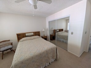 Ein Schlafzimmer mit einem Bett, einem Sessel, einem Nachttisch, einer Lampe, einem Spiegel und einem Deckenventilator.