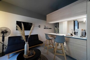 Ein modernes Wohnzimmer mit einem schwarzen Sofa, einer Essecke mit einem weißen Tisch und blauen Stühlen sowie einer Küche mit Einbauofen und Mikrowelle.