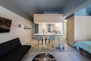 Ein modernes Wohnzimmer mit einer schwarzen Couch, einem Essbereich mit zwei Stühlen, einer Küche mit einer Kaffeemaschine und einem Bett mit blauer Decke.