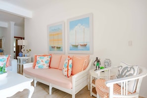 A cozy living room with a sofa, a small table, and a chair. There are two paintings on the wall, a white door, and a white ceiling.