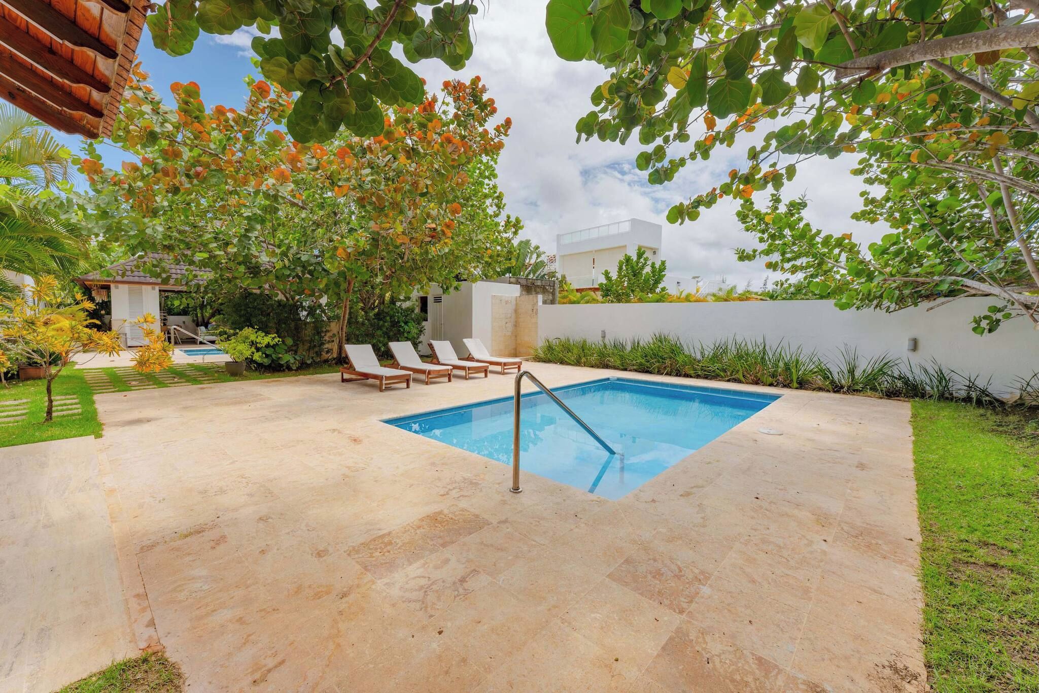 A swimming pool with a metal ladder, surrounded by a paved patio, trees, and a white wall.