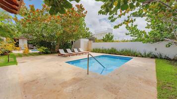 A swimming pool with a metal ladder, surrounded by a paved patio, trees, and a white wall.