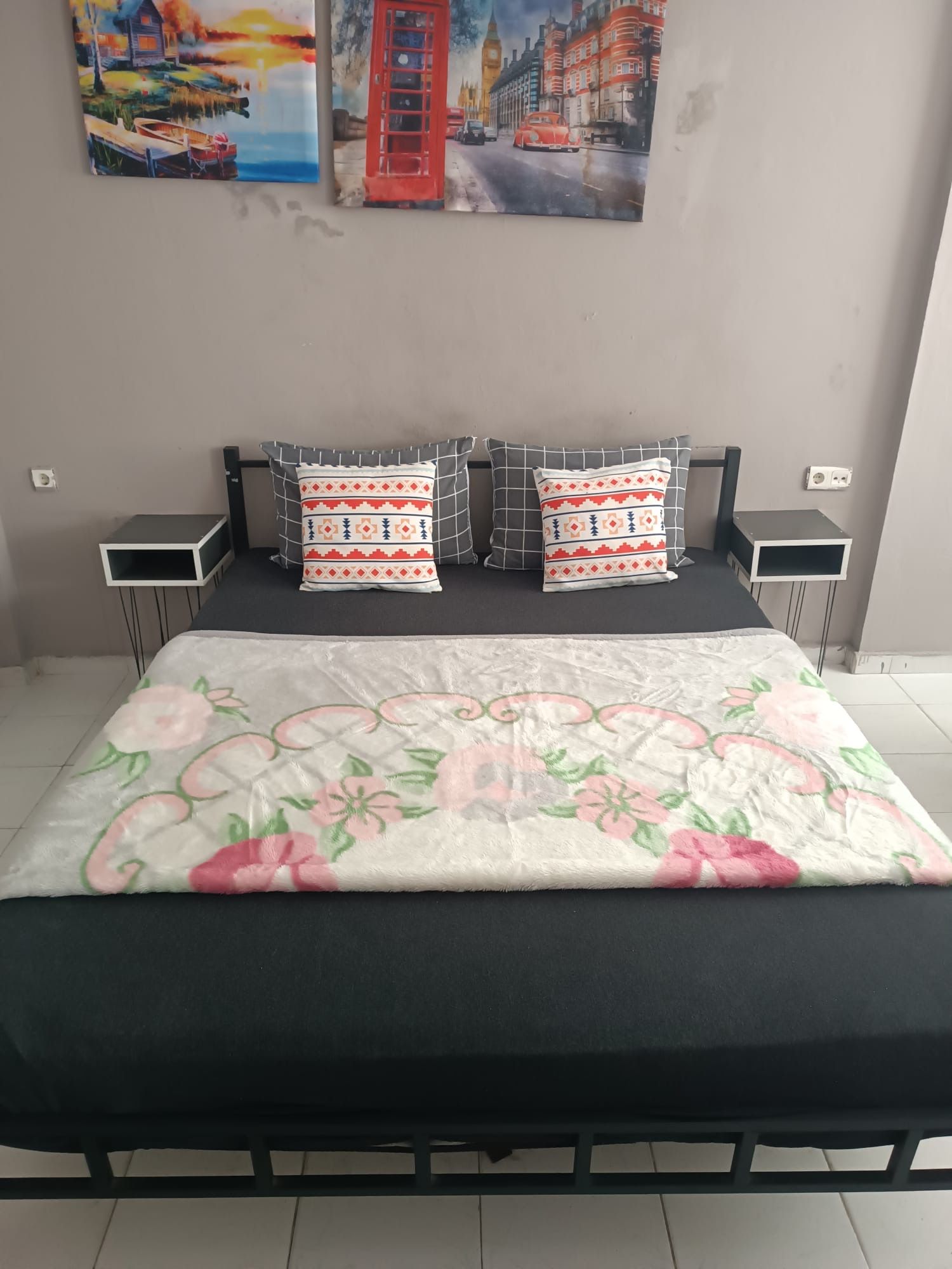 Una cama con colcha estampada de flores, dos almohadas con un diseño geométrico y un cuadro de un paisaje urbano sobre ella.